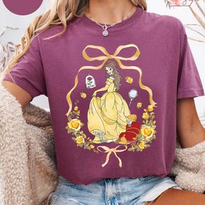 Peut inclure: T-shirt de couleur baie avec une illustration de dessin anim&eacute; d'une princesse en robe jaune, entour&eacute;e de roses jaunes et d'un n&oelig;ud dor&eacute;. Le motif comprend une rose dans une cloche et une petite figurine rouge.