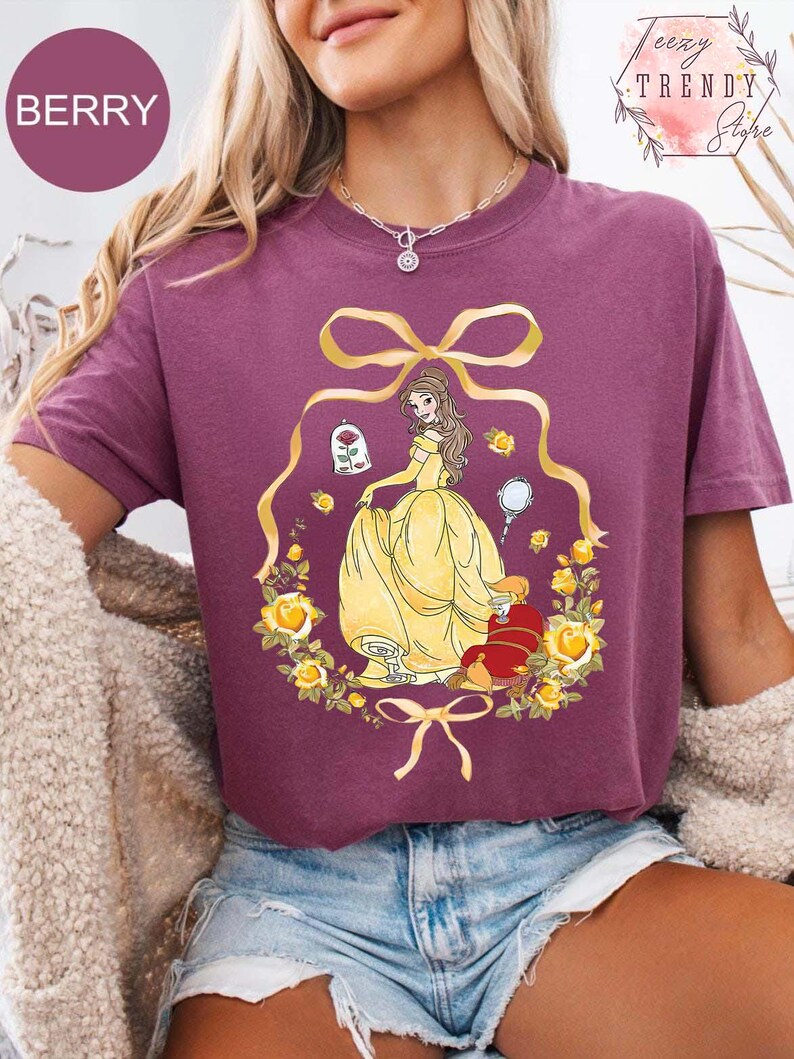 Peut inclure: T-shirt de couleur baie avec une illustration de dessin anim&eacute; d'une princesse en robe jaune, entour&eacute;e de roses jaunes et d'un n&oelig;ud dor&eacute;. Le motif comprend une rose dans une cloche et une petite figurine rouge.