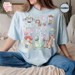 May include: Light blue t-shirt featuring a colorful graphic of characters from the animated film Toy Story. The design includes Buzz Lightyear, Woody, Jessie, Rex, Hamm, Slinky Dog, Bullseye, and the Little Green Men, with the text "You've Got a Friend in Me."