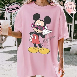 May include: Pink t-shirt featuring a graphic of Mickey Mouse wearing sunglasses. The cartoon character is in a relaxed pose, with the words "Mickey Mouse" in a script font. The shirt has the words "Comfort Colors Blossom" in a circular design.
