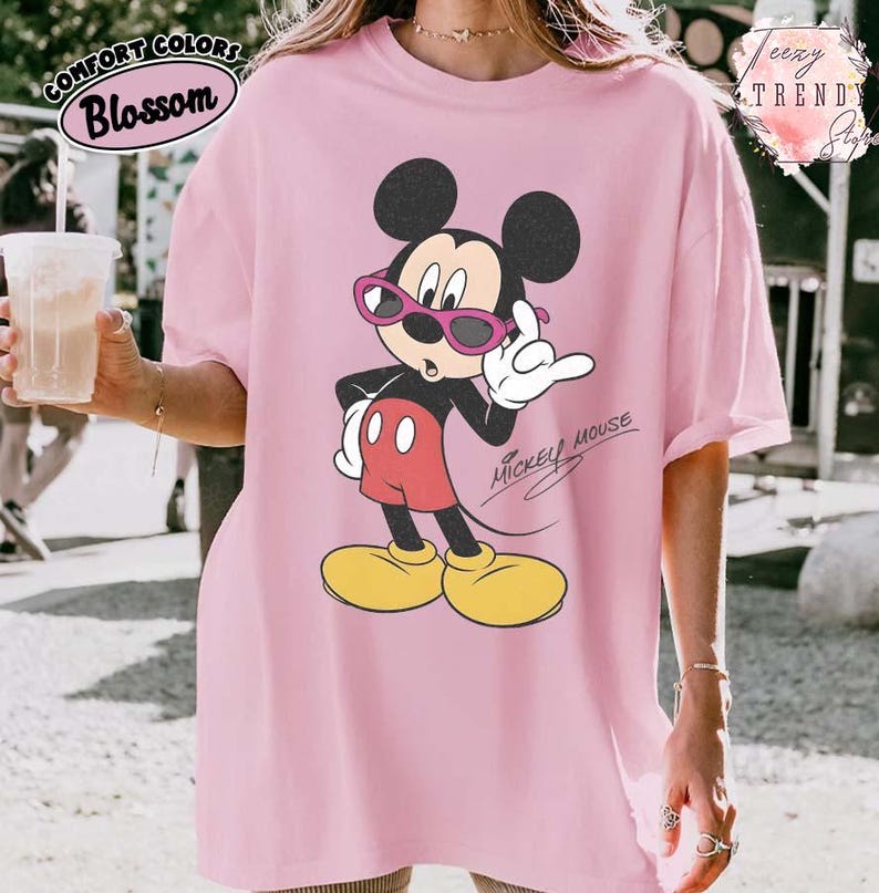 May include: Pink t-shirt featuring a graphic of Mickey Mouse wearing sunglasses. The cartoon character is in a relaxed pose, with the words "Mickey Mouse" in a script font. The shirt has the words "Comfort Colors Blossom" in a circular design.