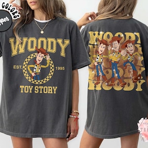 May include: Dark gray t-shirt featuring a Woody design from Toy Story. The front graphic includes the word "WOODY" in gold lettering, Woody cartoon, and "TOY STORY" text. The back of the shirt has a Woody design with the word "WOODY".