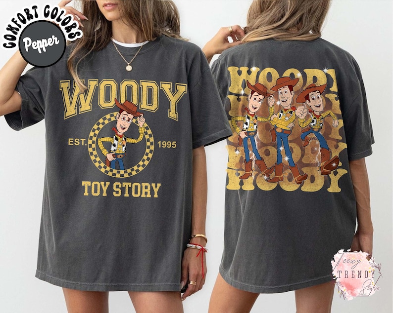 May include: Dark gray t-shirt featuring a Woody design from Toy Story. The front graphic includes the word "WOODY" in gold lettering, Woody cartoon, and "TOY STORY" text. The back of the shirt has a Woody design with the word "WOODY".