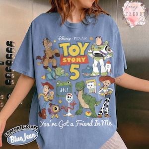 May include: A blue jean colored t-shirt featuring a graphic print of Toy Story characters including Woody, Buzz Lightyear, Jessie, Rex, Bullseye, and Forky. The design includes the text Toy Story 5 and You've Got a Friend In Me in white lettering.