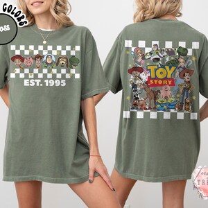 Op de afbeelding: Mosgroen t-shirt met personages uit de animatiefilm "Toy Story". De voorkant heeft een dambordontwerp met de tekst "EST. 1995". De achterkant toont een grotere afbeelding van de personages en de titel van de film.