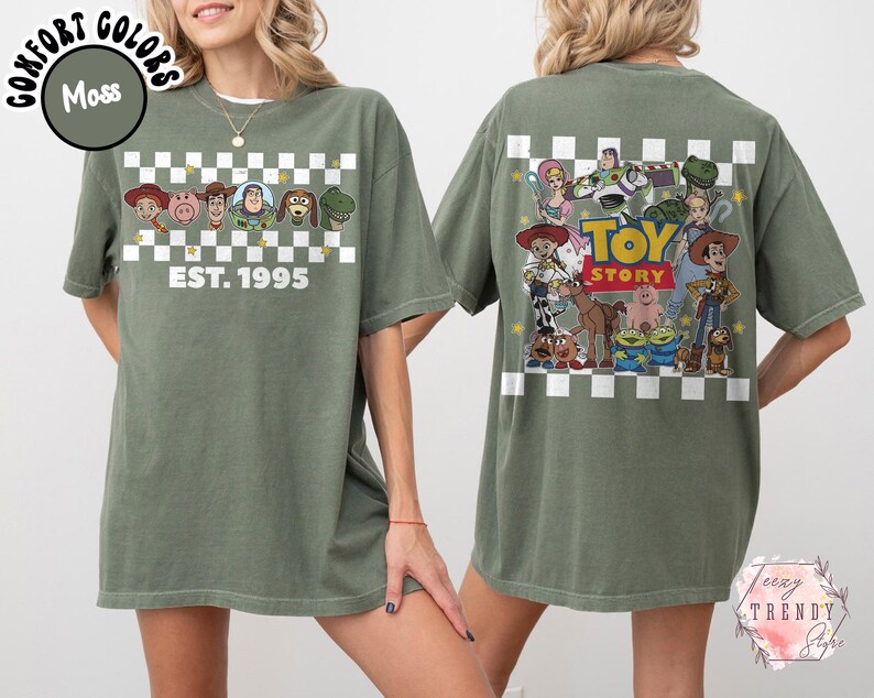 Op de afbeelding: Mosgroen t-shirt met personages uit de animatiefilm "Toy Story". De voorkant heeft een dambordontwerp met de tekst "EST. 1995". De achterkant toont een grotere afbeelding van de personages en de titel van de film.