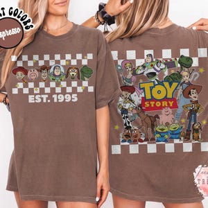 Op de afbeelding: Espresso-kleurig T-shirt met Toy Story-personages en de tekst "EST. 1995" op de voorkant. De achterkant van het shirt toont een grotere Toy Story-afbeelding met de woorden "TOY STORY". Het shirt heeft een dambordpatroon.