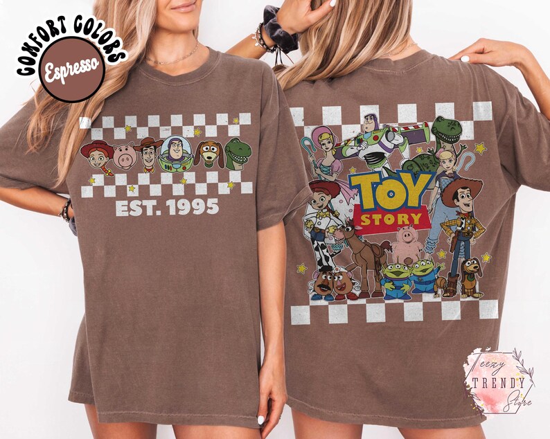 Op de afbeelding: Espresso-kleurig T-shirt met Toy Story-personages en de tekst "EST. 1995" op de voorkant. De achterkant van het shirt toont een grotere Toy Story-afbeelding met de woorden "TOY STORY". Het shirt heeft een dambordpatroon.