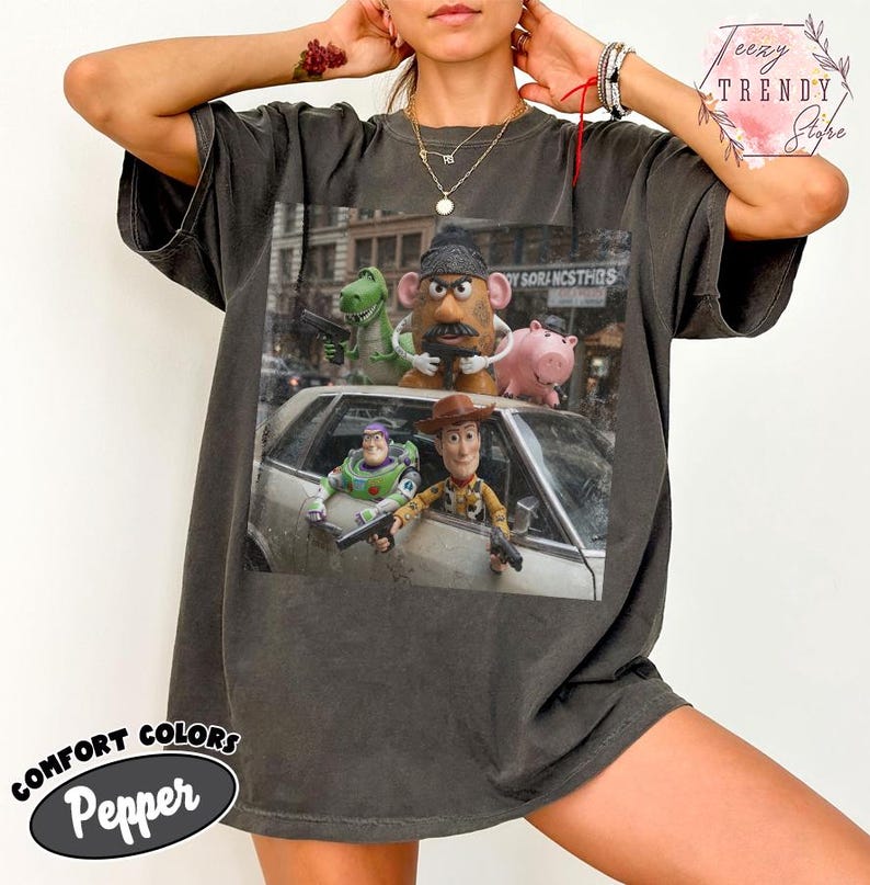 Puede incluir: Una camiseta de manga corta color gris carb&oacute;n con un estampado gr&aacute;fico de personajes de Toy Story, incluyendo a Woody, Buzz Lightyear, el Sr. Patata, Rex y Hamm, posando con pistolas de juguete alrededor de un coche plateado en la ciudad.