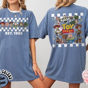Op de afbeelding: Blauwe jeanskleurige t-shirt met personages uit de animatiefilm "Toy Story". De voorkant toont personages en de tekst "EST. 1995" boven een dambordpatroon. De achterkant toont een grotere afbeelding van de personages van de film met de woorden "Toy Story".