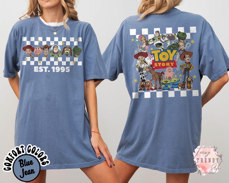 Op de afbeelding: Blauwe jeanskleurige t-shirt met personages uit de animatiefilm "Toy Story". De voorkant toont personages en de tekst "EST. 1995" boven een dambordpatroon. De achterkant toont een grotere afbeelding van de personages van de film met de woorden "Toy Story".