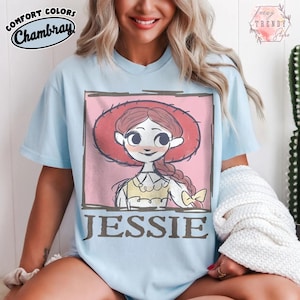 May include: Pale blue t-shirt with a cartoon illustration of Jessie from Toy Story. The graphic is set within a pink square and includes the name "JESSIE" in brown letters. The shirt has the words "COMFORT COLORS Chambray" printed on the top left.