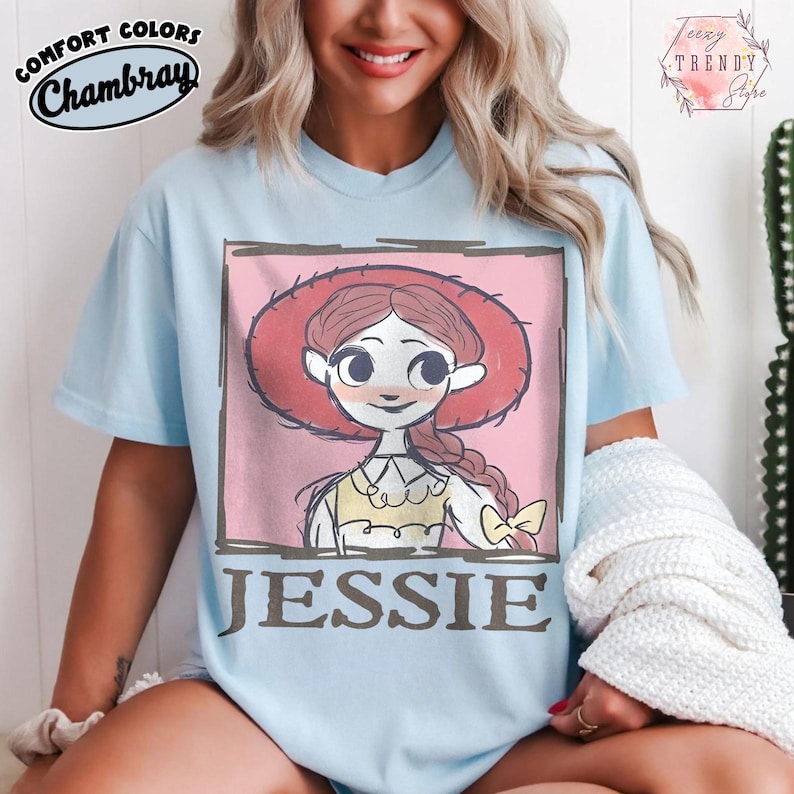 May include: Pale blue t-shirt with a cartoon illustration of Jessie from Toy Story. The graphic is set within a pink square and includes the name "JESSIE" in brown letters. The shirt has the words "COMFORT COLORS Chambray" printed on the top left.