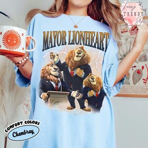 Puede incluir: Camiseta azul claro con un gr&aacute;fico de tres leones con trajes y el texto "MAYOR LIONHEART". La camiseta tambi&eacute;n tiene el texto "COMFORT COLORS Chambray". En la imagen se sostiene una taza blanca y naranja con un dise&ntilde;o de sol.