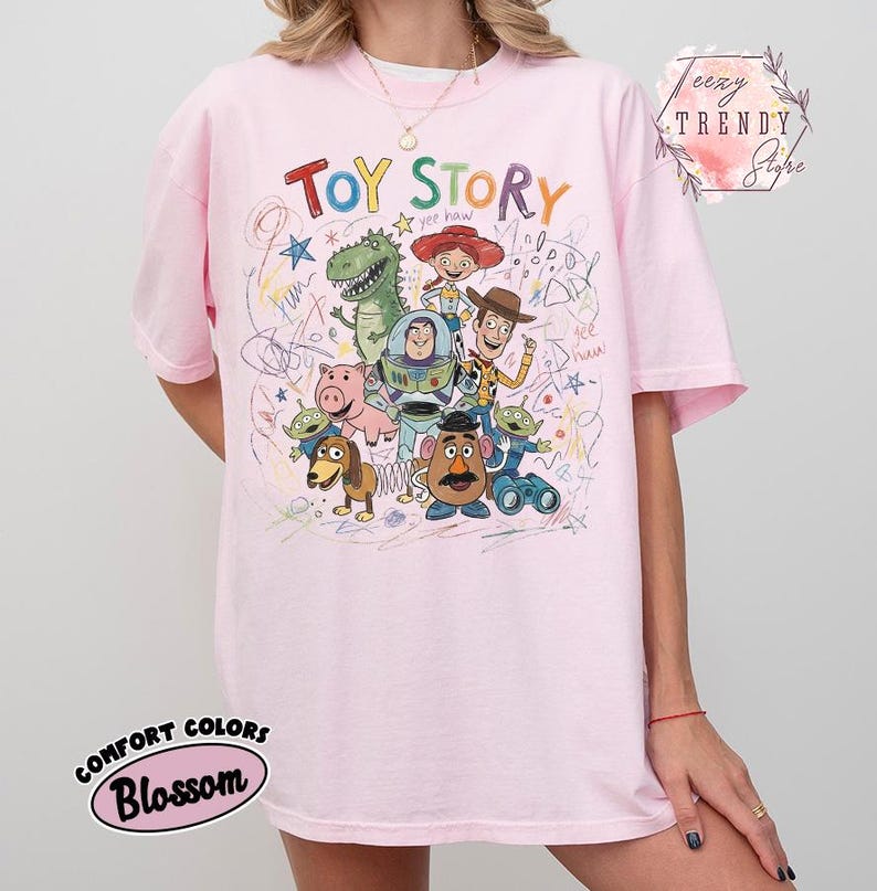 May include: Pink t-shirt featuring a colorful graphic of Toy Story characters, including Woody, Buzz Lightyear, and Rex. The words "Toy Story" are written in a playful font above the characters. The shirt is a Comfort Colors Blossom.