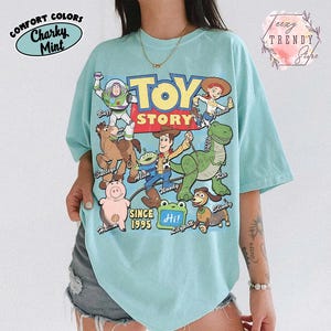 May include: A light mint green t-shirt featuring a colorful graphic print of Toy Story characters including Buzz Lightyear, Woody, Jessie, Bullseye, Rex, Hamm, Slinky, and an Alien. The text reads Toy Story, Since 1995, and Hi! Lilypad.