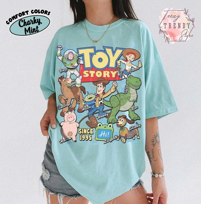 May include: A light mint green t-shirt featuring a colorful graphic print of Toy Story characters including Buzz Lightyear, Woody, Jessie, Bullseye, Rex, Hamm, Slinky, and an Alien. The text reads Toy Story, Since 1995, and Hi! Lilypad.