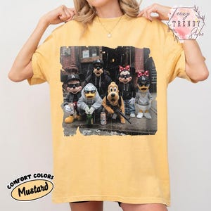 May include: A mustard yellow t-shirt featuring a graphic print of five cartoon characters wearing black baseball caps and sunglasses. The characters are dressed in casual streetwear, including hoodies and jackets, while standing together in an urban setting.