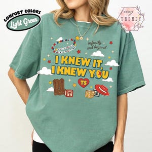 Puede incluir: Una camiseta verde claro con un estampado gr&aacute;fico que incluye el texto I KNEW IT, I KNEW YOU, infinity and beyond y YOU'VE GOT A FRIEND IN ME. El dise&ntilde;o presenta ilustraciones de botas vaqueras marrones, un sombrero rojo, bloques y estrellas.