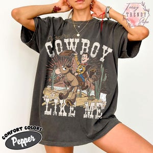 May include: A charcoal gray short-sleeve t-shirt featuring a graphic print of a cartoon cowboy riding a horse in a desert landscape. The text reads You're a COWBOY LIKE ME. The design includes cacti, a sunburst, and rocky terrain in muted tones.