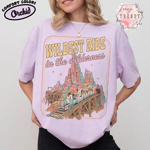 Może przedstawiać: Koszulka Comfort Colors w kolorze orchidei z grafiką przedstawiającą pociąg z Myszką Miki i Minnie oraz innymi postaciami na torze przez pasmo g&oacute;rskie. Tekst brzmi "Wildest Ride in the wilderness."