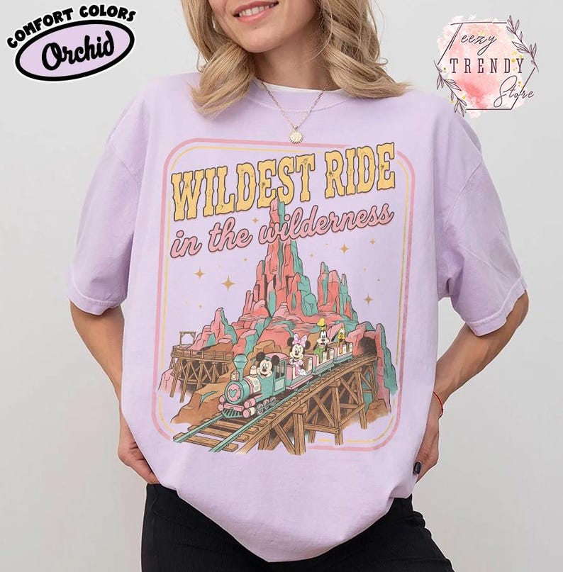 Może przedstawiać: Koszulka Comfort Colors w kolorze orchidei z grafiką przedstawiającą pociąg z Myszką Miki i Minnie oraz innymi postaciami na torze przez pasmo g&oacute;rskie. Tekst brzmi "Wildest Ride in the wilderness."