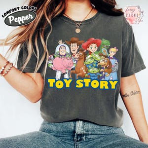 Puede incluir: Camiseta Comfort Colors gris oscuro con un gr&aacute;fico de personajes de Toy Story, incluyendo a Woody, Buzz Lightyear y otros. Las palabras "TOY STORY" est&aacute;n impresas en amarillo y azul. La camiseta tiene un corte relajado.