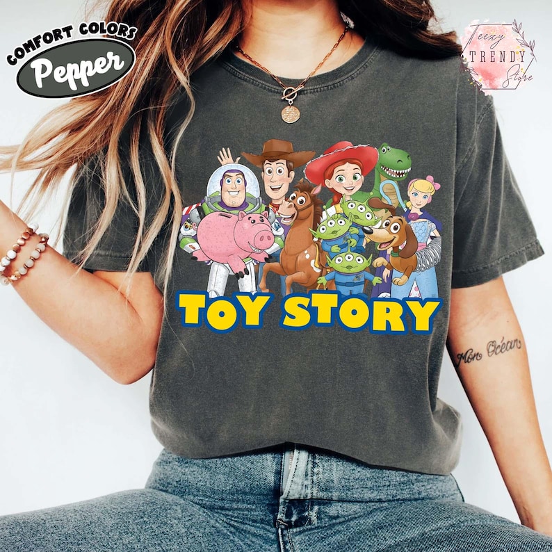 Puede incluir: Camiseta Comfort Colors gris oscuro con un gr&aacute;fico de personajes de Toy Story, incluyendo a Woody, Buzz Lightyear y otros. Las palabras "TOY STORY" est&aacute;n impresas en amarillo y azul. La camiseta tiene un corte relajado.