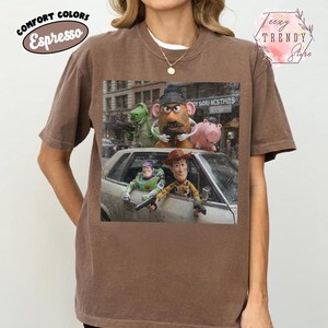 Puede incluir: Una camiseta de manga corta color marr&oacute;n espresso con un estampado gr&aacute;fico de personajes de Toy Story, incluyendo a Woody, Buzz Lightyear, Mr. Potato Head, Rex y Hamm. Los personajes aparecen en una calle con un coche plateado.