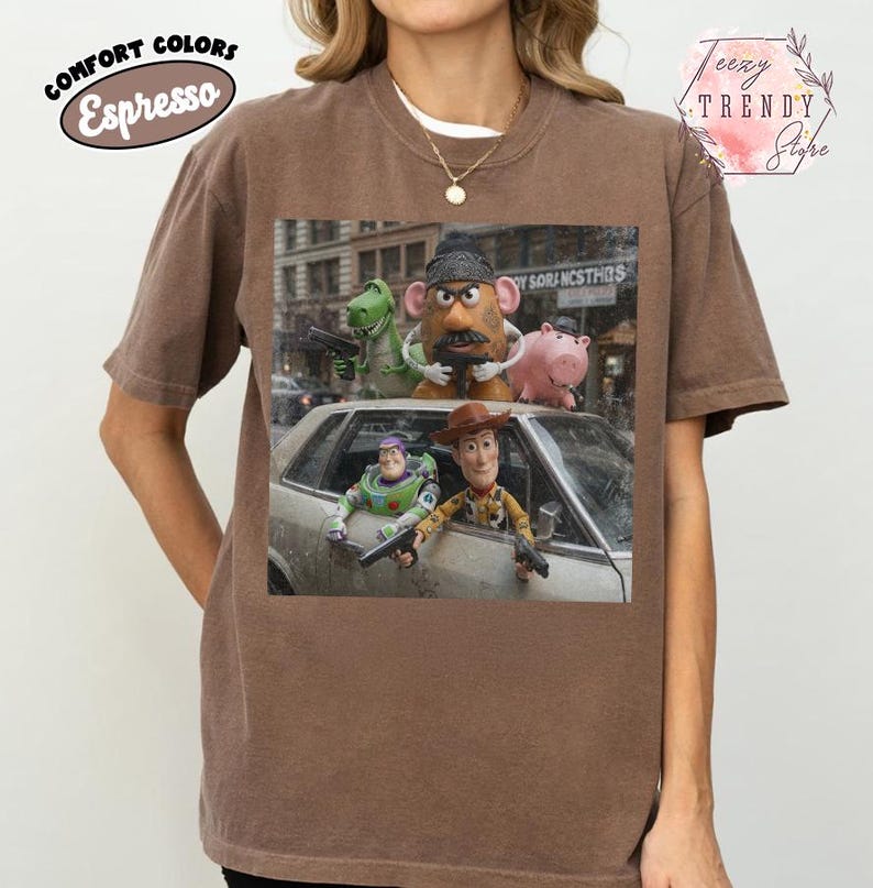 Puede incluir: Una camiseta de manga corta color marr&oacute;n espresso con un estampado gr&aacute;fico de personajes de Toy Story, incluyendo a Woody, Buzz Lightyear, Mr. Potato Head, Rex y Hamm. Los personajes aparecen en una calle con un coche plateado.