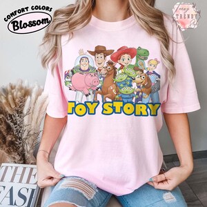 Puede incluir: Camiseta rosa claro con un estampado colorido de personajes de la pel&iacute;cula "Toy Story". El dise&ntilde;o incluye a Buzz Lightyear, Woody, Jessie y otros personajes. Las palabras "TOY STORY" est&aacute;n impresas en amarillo con un contorno azul. La camiseta tiene los logotipos "Comfort Colors" y "Blossom".