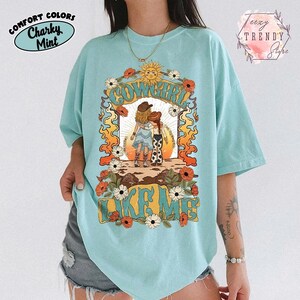 May include: A light mint green t-shirt featuring a graphic print of two figures in cowboy hats standing together. The text "COWGIRL LIKE ME" is printed in a bold, stylized font. The design includes a sun, orange flames, and white and red flowers.