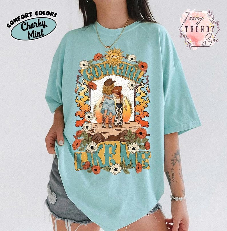 May include: A light mint green t-shirt featuring a graphic print of two figures in cowboy hats standing together. The text "COWGIRL LIKE ME" is printed in a bold, stylized font. The design includes a sun, orange flames, and white and red flowers.