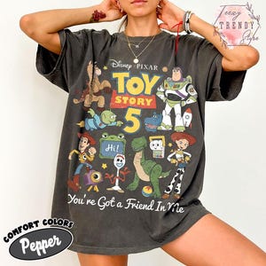 May include: A charcoal gray short-sleeve t-shirt featuring a graphic print of Disney Pixar Toy Story characters including Buzz Lightyear, Woody, Jessie, Rex, and Forky. The text reads Toy Story 5 and You're Got a Friend In Me.
