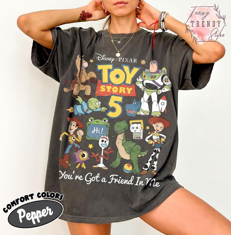 May include: A charcoal gray short-sleeve t-shirt featuring a graphic print of Disney Pixar Toy Story characters including Buzz Lightyear, Woody, Jessie, Rex, and Forky. The text reads Toy Story 5 and You're Got a Friend In Me.