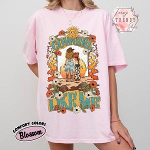 May include: A light pink t-shirt featuring a graphic print of two figures in cowboy hats and boots standing under a sun. The text reads COWGIRL LIKE ME. The design is framed by orange flames and white and orange flowers on a light pink background.