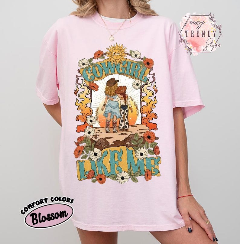 May include: A light pink t-shirt featuring a graphic print of two figures in cowboy hats and boots standing under a sun. The text reads COWGIRL LIKE ME. The design is framed by orange flames and white and orange flowers on a light pink background.