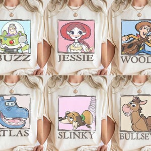May include: Cream-coloured t-shirts with cartoon character portraits in rectangular frames. Characters include Buzz Lightyear, Jessie, Woody, Atlas, Slinky, and Bullseye, each with their name below. The designs are in a cartoon style.