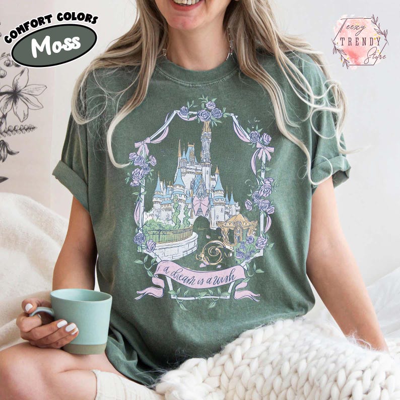 Puede incluir: Camiseta verde musgo con un gr&aacute;fico de un castillo de cuento de hadas, rodeado por una corona floral y una pancarta que dice "a dream is a wish". La camiseta tiene las mangas enrolladas. Las palabras "Comfort Colors" y "Moss" est&aacute;n impresas en la parte superior izquierda.