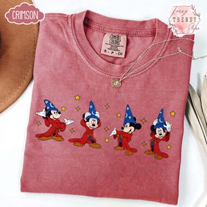 May include: A dusty rose Comfort Colors t-shirt featuring a graphic of Mickey Mouse in a sorcerer's outfit. The design includes Mickey in a blue hat and red robe, with yellow stars. A silver necklace with a pendant is also visible.