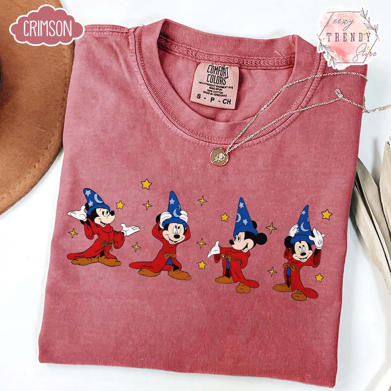 May include: A dusty rose Comfort Colors t-shirt featuring a graphic of Mickey Mouse in a sorcerer's outfit. The design includes Mickey in a blue hat and red robe, with yellow stars. A silver necklace with a pendant is also visible.
