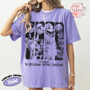 May include: A violet short-sleeve t-shirt featuring a black graphic print of characters from Tim Burton's The Nightmare Before Christmas. The design consists of four vertical panels displaying Jack Skellington, Sally, Oogie Boogie, and Lock, Shock, and Barrel.