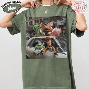 Puede incluir: Una camiseta de manga corta color verde musgo con un estampado gr&aacute;fico de personajes de Toy Story, incluidos Woody, Buzz Lightyear, Rex, el Sr. Patata y Hamm, representados en un entorno urbano con pistolas de juguete.