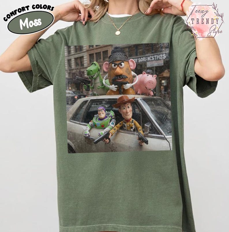 Puede incluir: Una camiseta de manga corta color verde musgo con un estampado gr&aacute;fico de personajes de Toy Story, incluidos Woody, Buzz Lightyear, Rex, el Sr. Patata y Hamm, representados en un entorno urbano con pistolas de juguete.