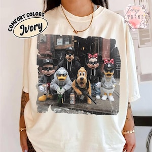 May include: An ivory t-shirt featuring a graphic print of five cartoon characters wearing hoodies and baseball caps. The characters are standing in a group, with some holding beverage cans and one holding a rifle. The print has a distressed texture.