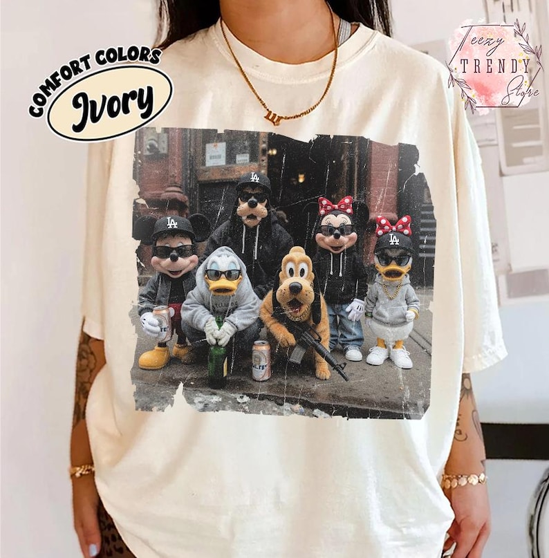May include: An ivory t-shirt featuring a graphic print of five cartoon characters wearing hoodies and baseball caps. The characters are standing in a group, with some holding beverage cans and one holding a rifle. The print has a distressed texture.
