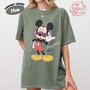 May include: Moss green t-shirt featuring a graphic of Mickey Mouse wearing sunglasses. The cartoon character is in a pose with one hand up. The words "Mickey Mouse" are written in cursive. The shirt is part of the Comfort Colors collection.