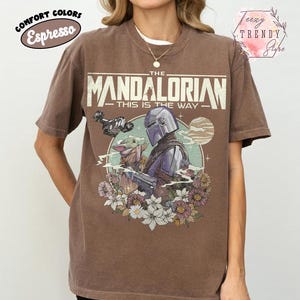 May include: Espresso-coloured t-shirt with a graphic of The Mandalorian and Grogu surrounded by flowers. The text "THE MANDALORIAN - THIS IS THE WAY" is printed across the top. The shirt has short sleeves and a crew neck.