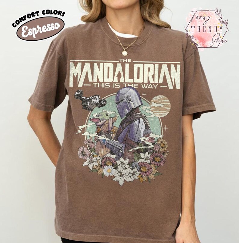 May include: Espresso-coloured t-shirt with a graphic of The Mandalorian and Grogu surrounded by flowers. The text "THE MANDALORIAN - THIS IS THE WAY" is printed across the top. The shirt has short sleeves and a crew neck.