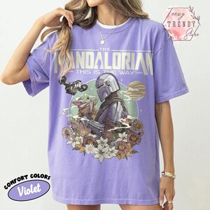 May include: A violet Comfort Colors t-shirt with a graphic of The Mandalorian and Grogu, surrounded by flowers. The design includes the text "THE MANDALORIAN - THIS IS THE WAY". The shirt has a relaxed fit.